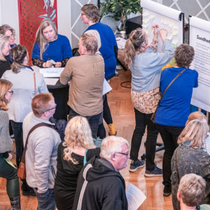 Mange mennesker samlet i et stort lokale. Nogle står og snakker ved caféborde, mens andre hænger post its på et lærred.