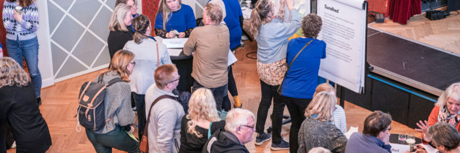 Mange mennesker samlet i et stort lokale. Nogle står og snakker ved caféborde, mens andre hænger post its på et lærred.